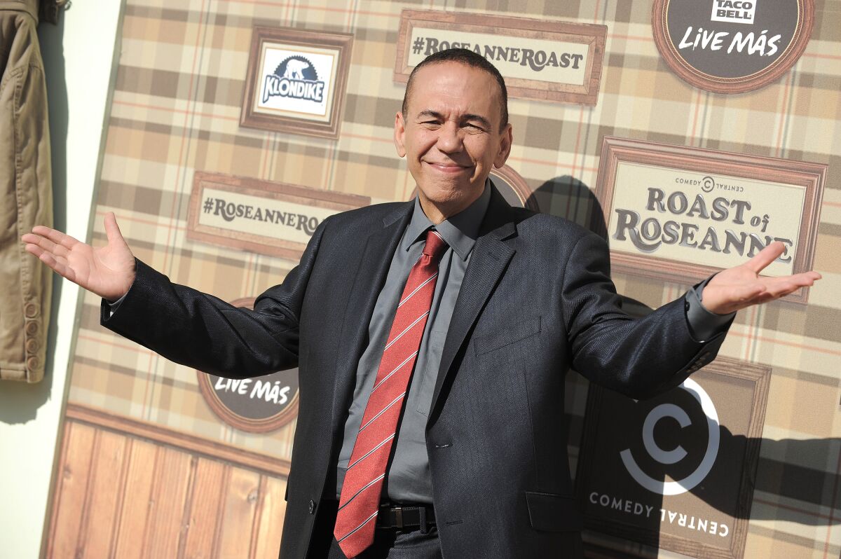 A man in a suit smirks and squints in the sun with his arms wide open.
