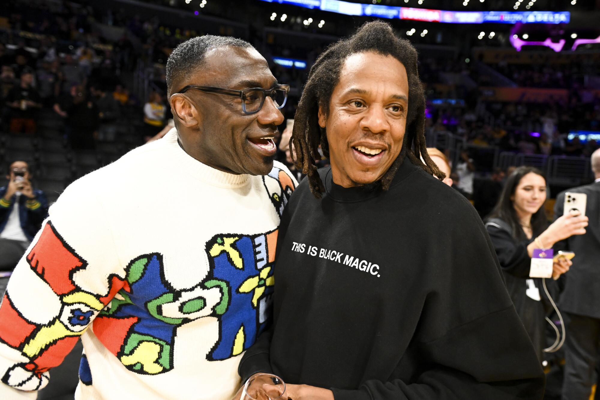Shannon Sharpe in a sweater and glasses talking and standing close to Jay-Z in a 'This Is Black Magic' sweatshirt