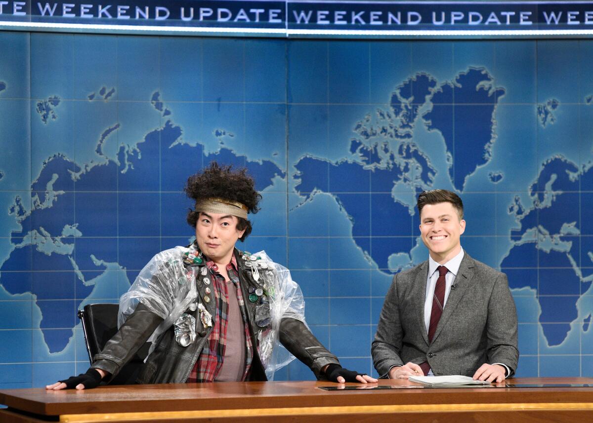 Two men, one in costume, sit behind a news desk set  