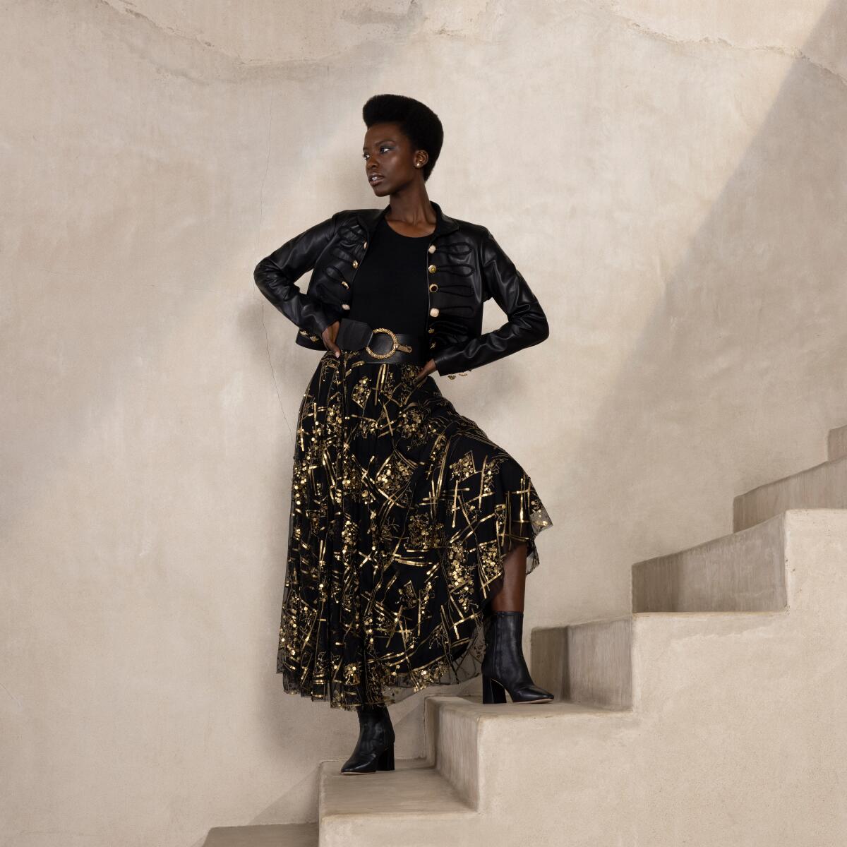 A model posing on a staircase in a sparkly gold and black skirt and short jacket