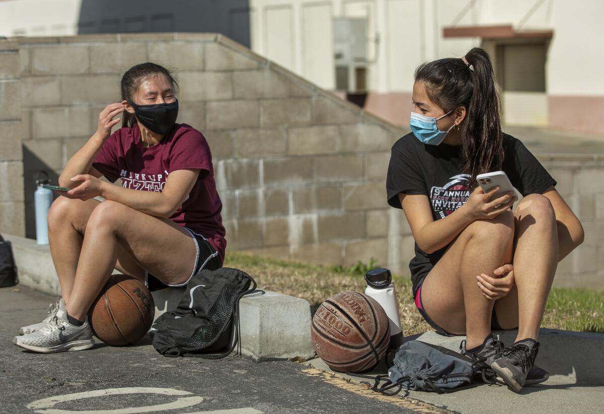 Two masked girls sit outside a school with basketballs.