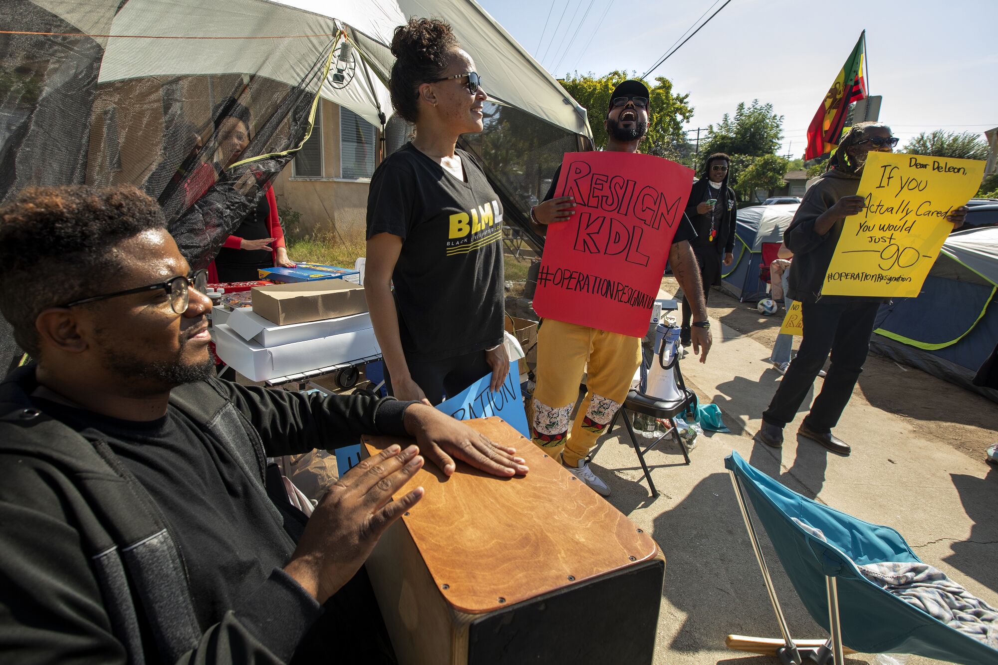 Protesters at a tent encampment hold signs reading "Resign KDL" and "If You Actually Cared, You Would Just Go."
