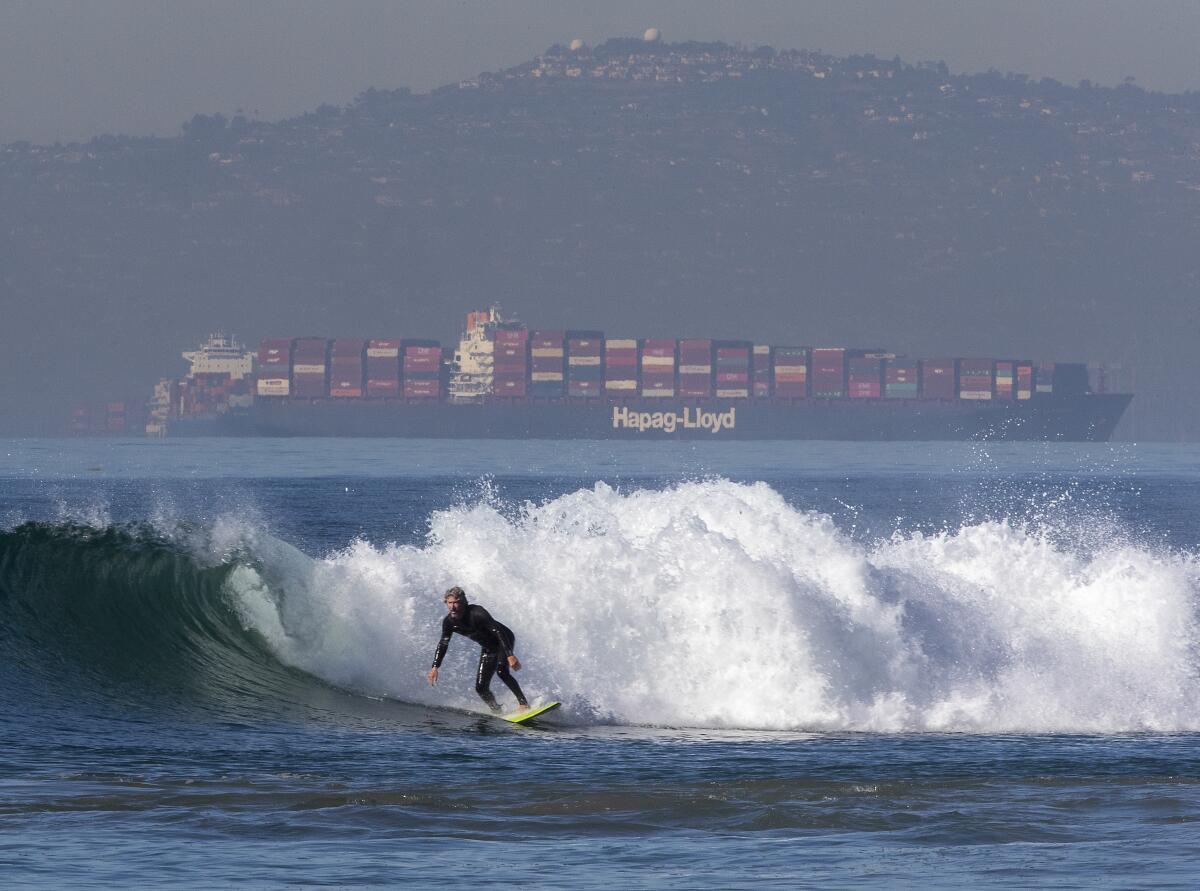 A surfer rides a wave as a large cargo ship floats in the distance