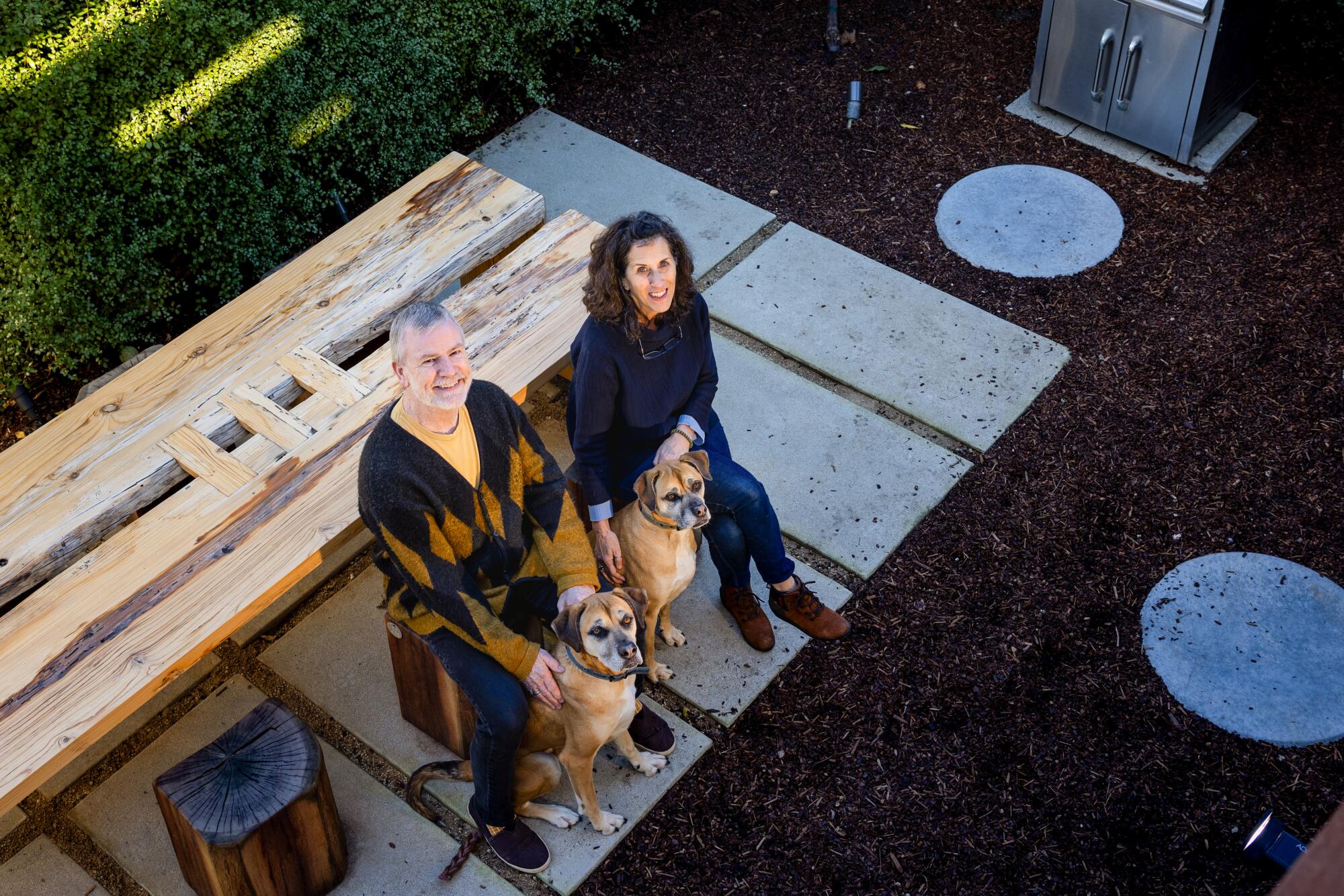 Architects Jefferson Schierbeek and Su Addison look up while sitting at a backyard table with two dogs at their feet.