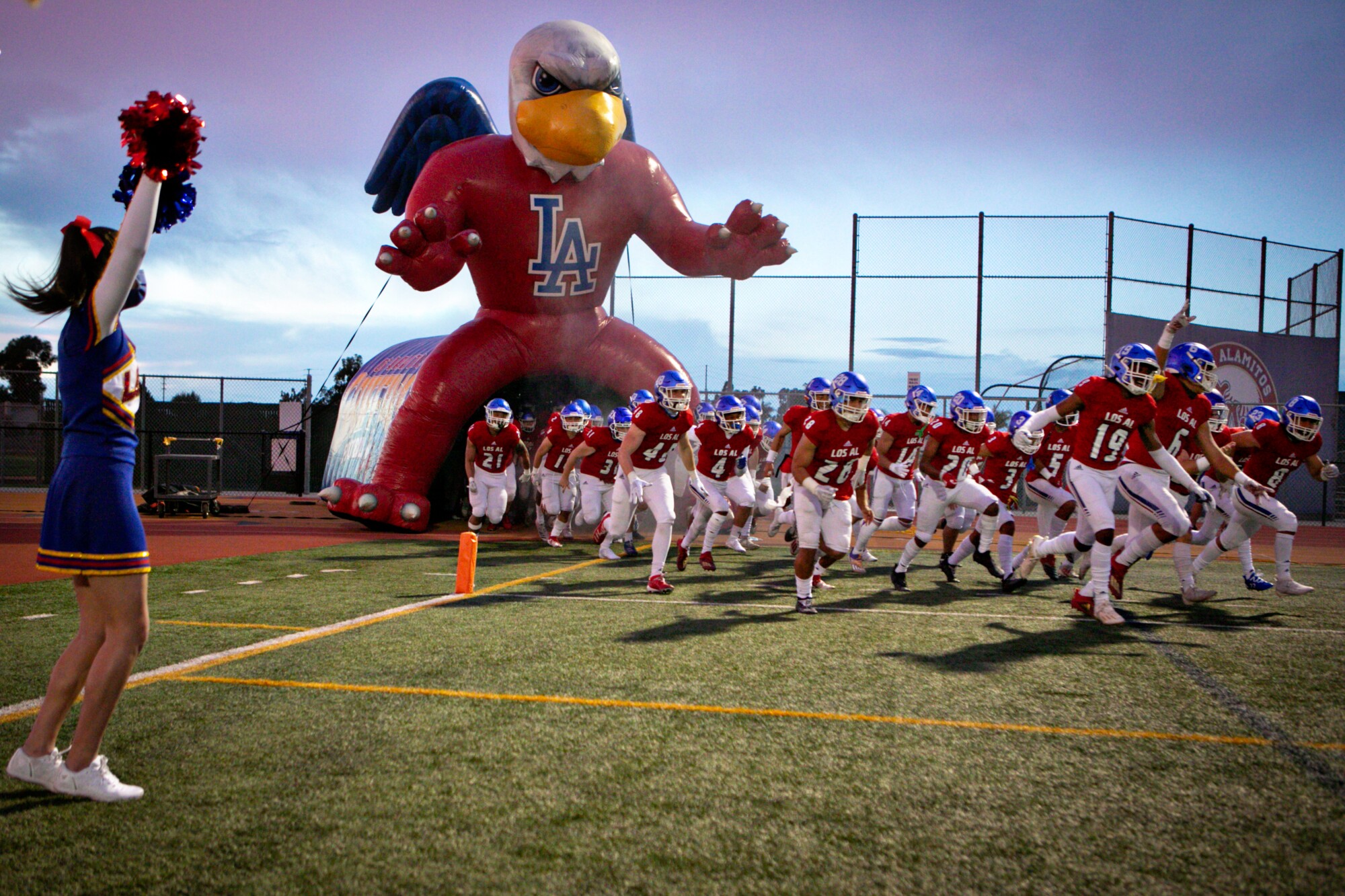The Los Alamitos football team enters the field through an inflatable tunnel with a cheerleader cheering nearby.