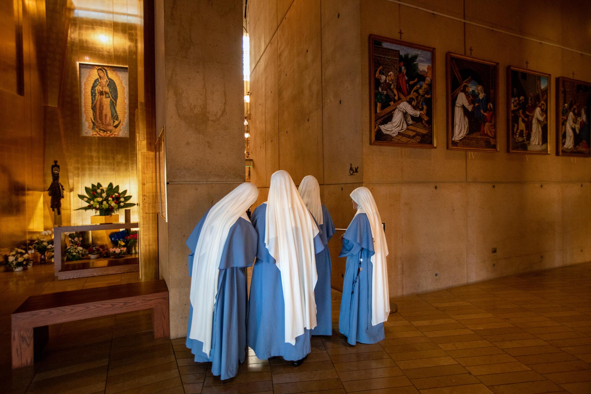Four nuns in blue and white habits face away in a huddle as they peak into the main hall of a modern stone church.