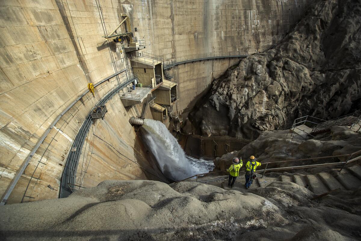 two workers are dwarfed by the towering wall of a dam. 