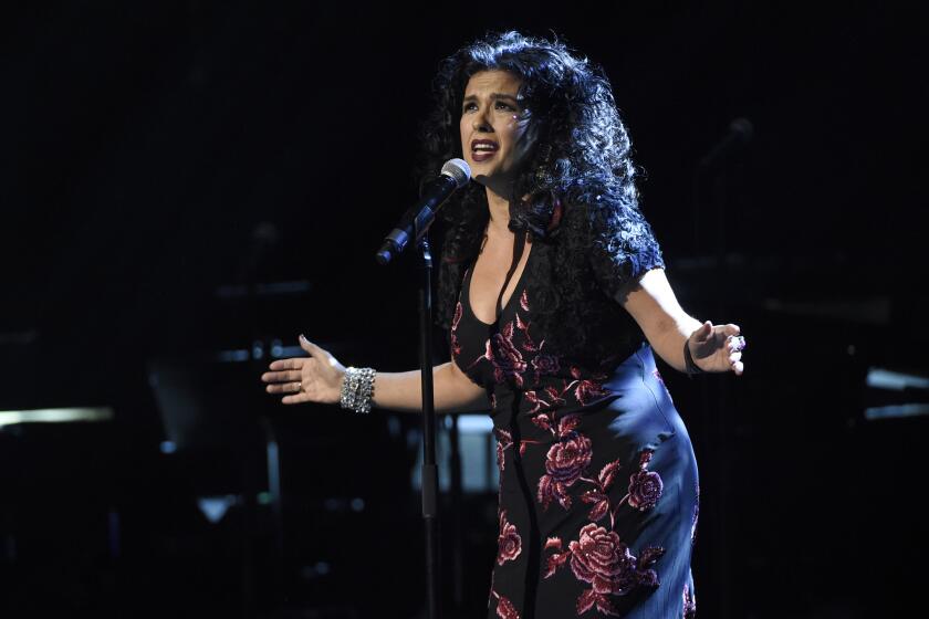 Rebekah Del Rio leaning into a microphone on a dimly lit stage with her arms outspread