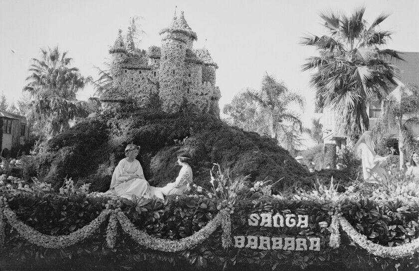Two young women in white gowns are seated on a float amid greenery, with a small castle made of flowers behind them.