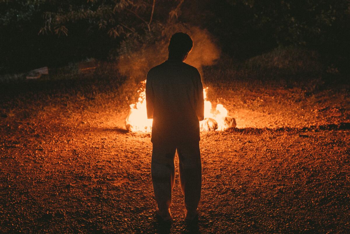 A figure observes a bonfire in the dark.