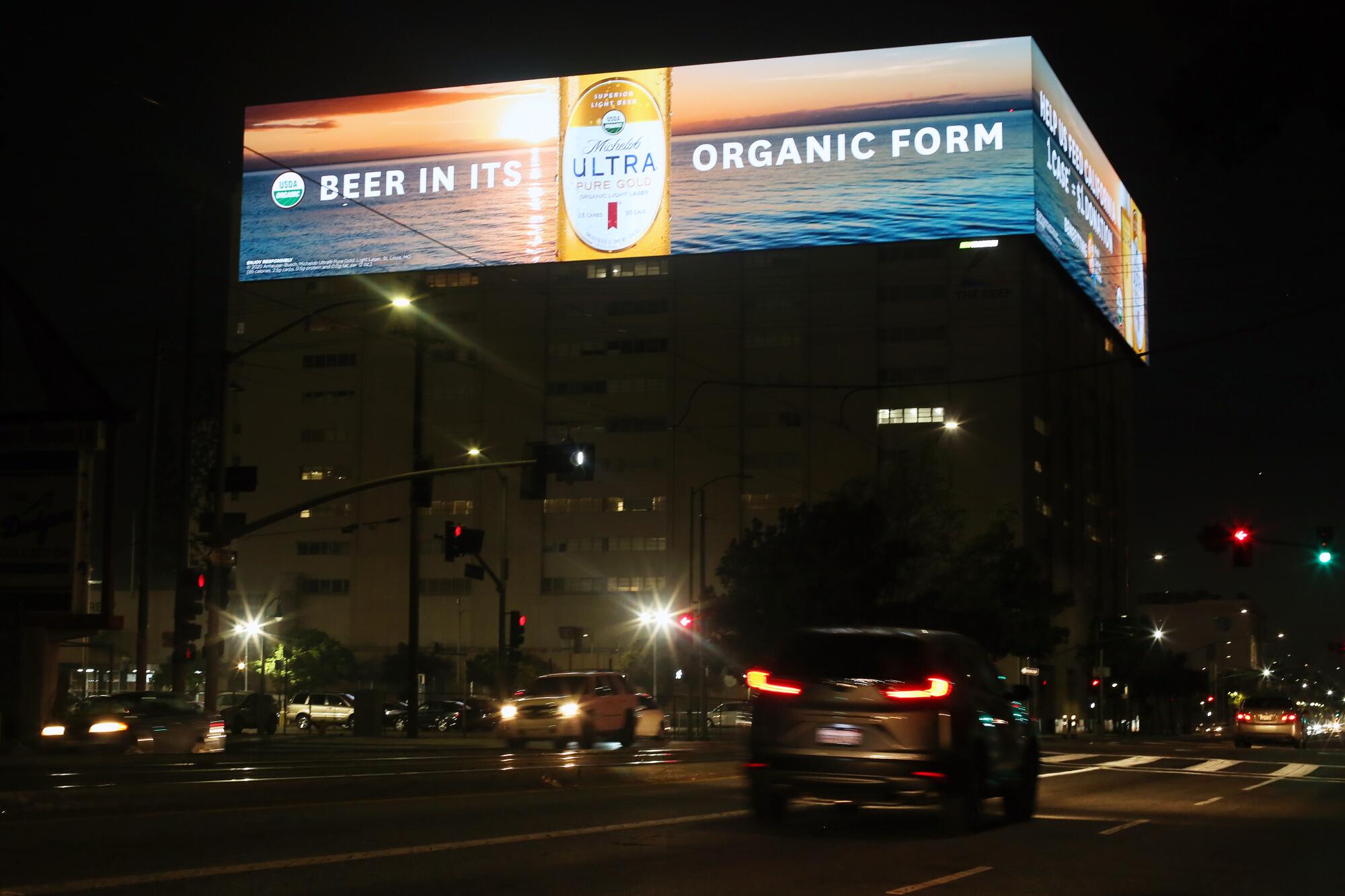 Cars on a nighttime city street pass below brightly lighted electronic billboards advertising beer