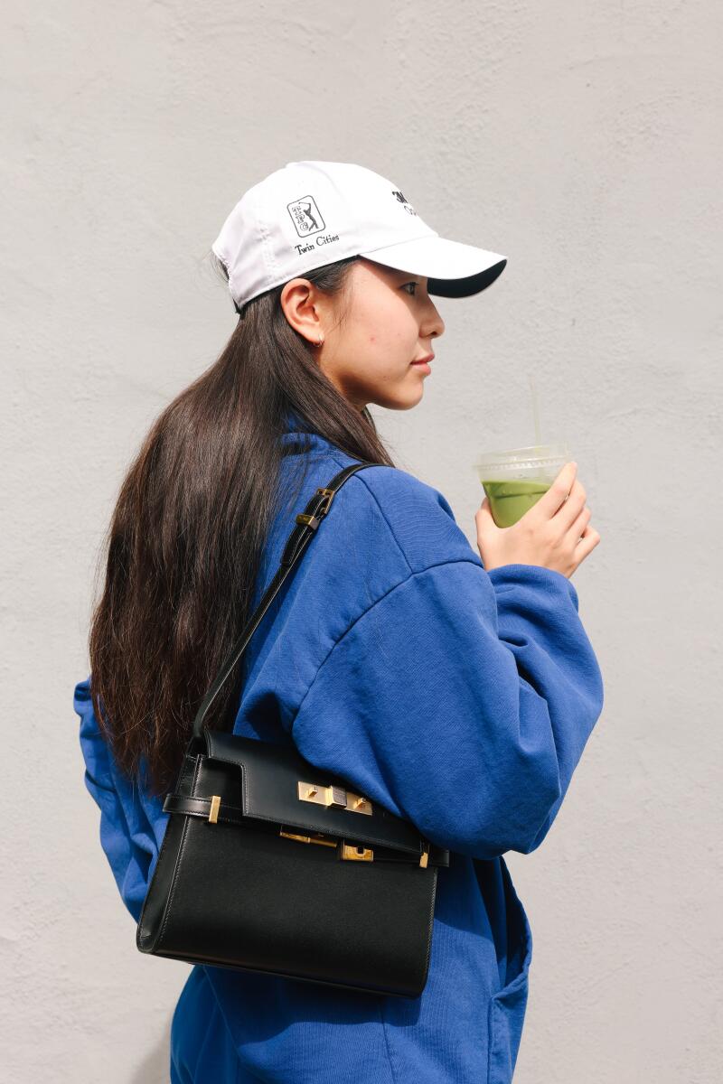 A woman in a white baseball cap and blue jumpsuit holds a matcha drink