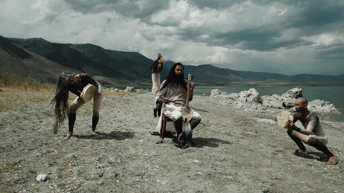 A film still shows three musicians in smoky-colored clothes playing instruments on the shore of a rocky lake