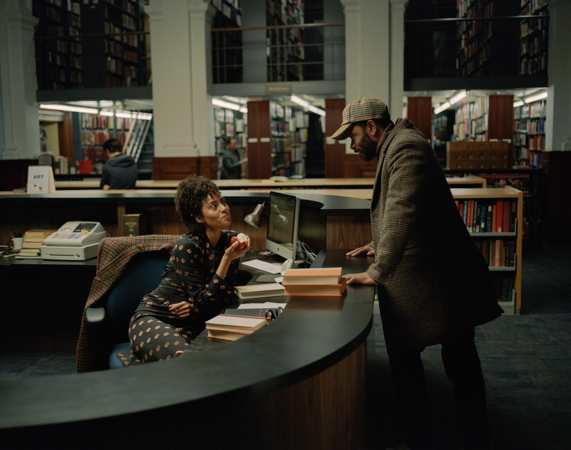 A librarian sits behind a counter holding an apple while a man stands on the other side speaking to her.