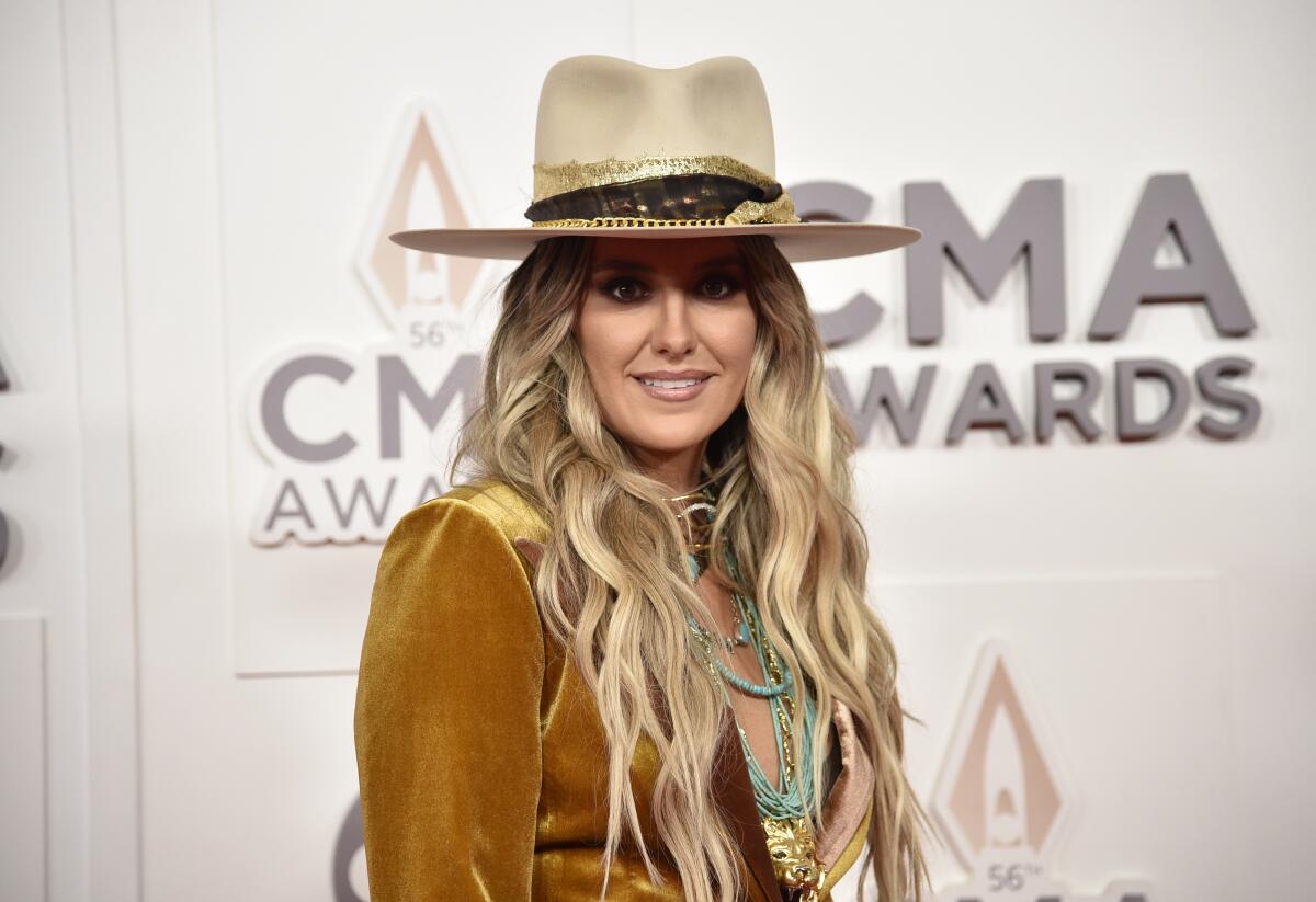 A woman with long blond hair wears a tall, wide-brim hat and stands in front of a CMA Awards backdrop
