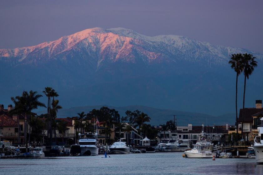Huntington Beach, CA - February 23: A person on a stand-up paddle board takes a scenic cruise as the setting sun illuminates the top of recently snow-capped San Gabriel mountains in Huntington Harbour in Huntington Beach Friday, Feb. 23, 2024. (Allen J. Schaben / Los Angeles Times)