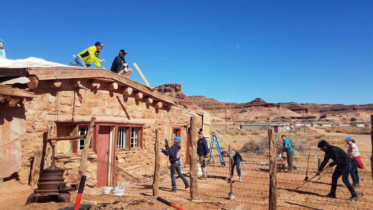Two people work on the roof of an old brick building and six stand on the dirt in front of it