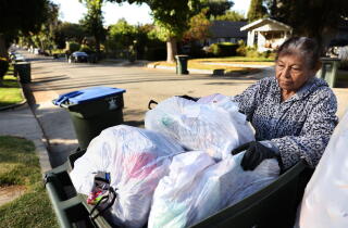 A woman wearing gloves looks over bags of garbage in a full trash bin on the curb of a sunny residential street.