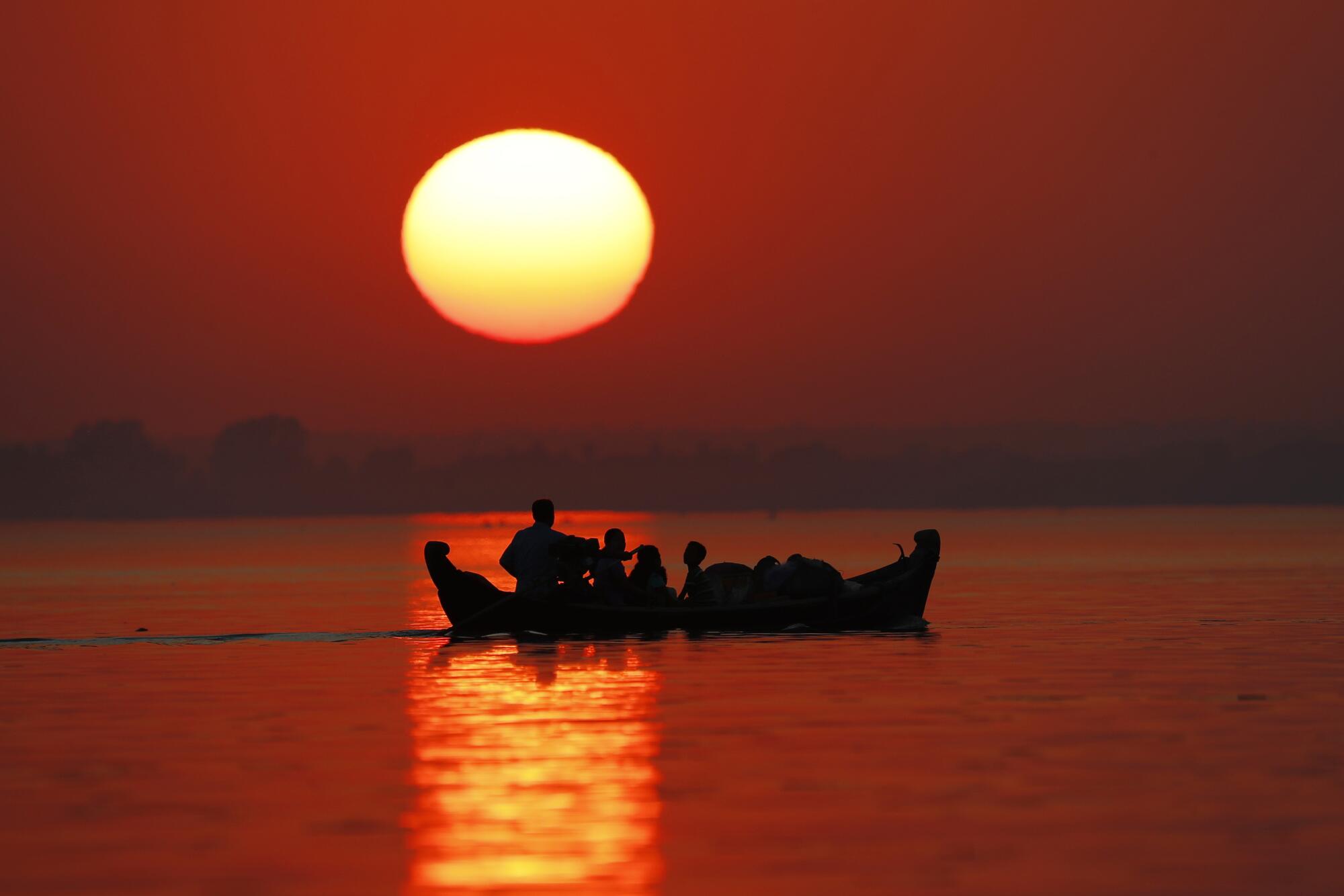 People and a small boat are in silhouette on a river, with the sun behind them, large and low on the horizon.