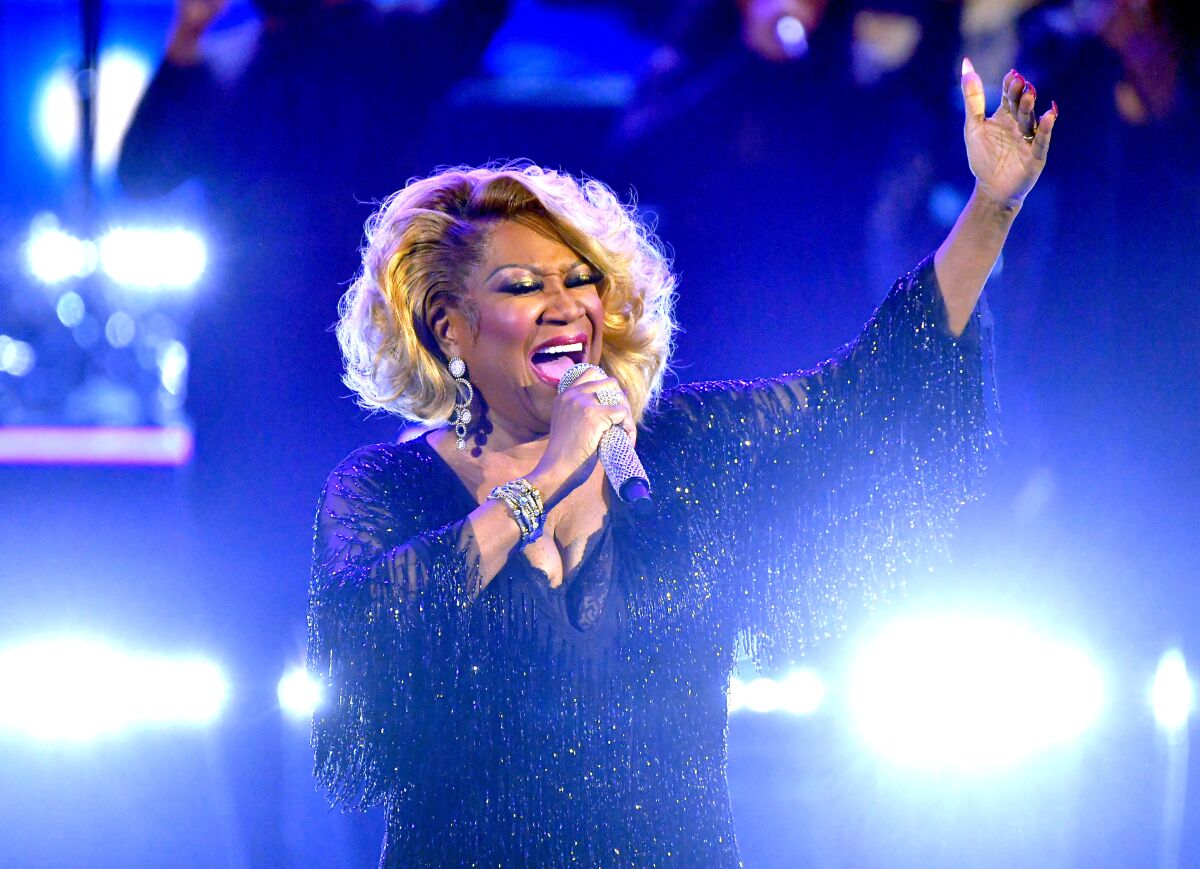 Patti LaBelle singing into a microphone on stage with her hand raised, wearing a sparkly dress with tasles