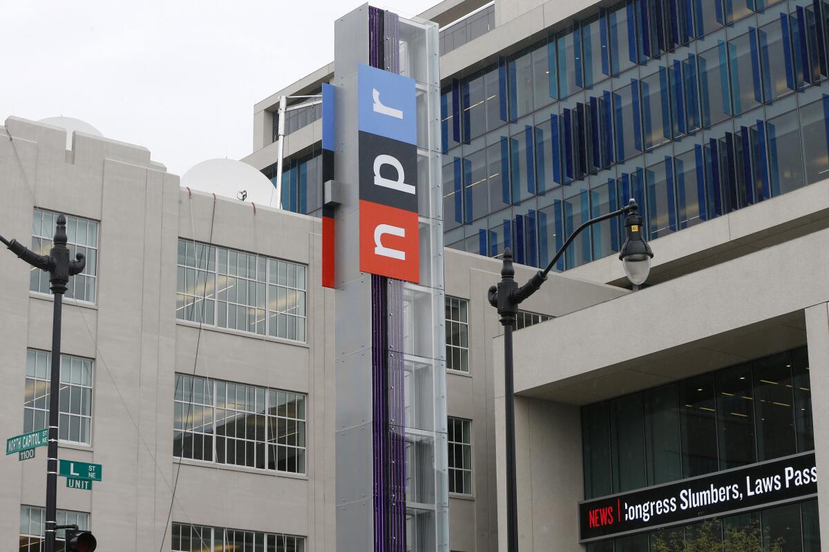 An NPR sign on a column outside gray buildings