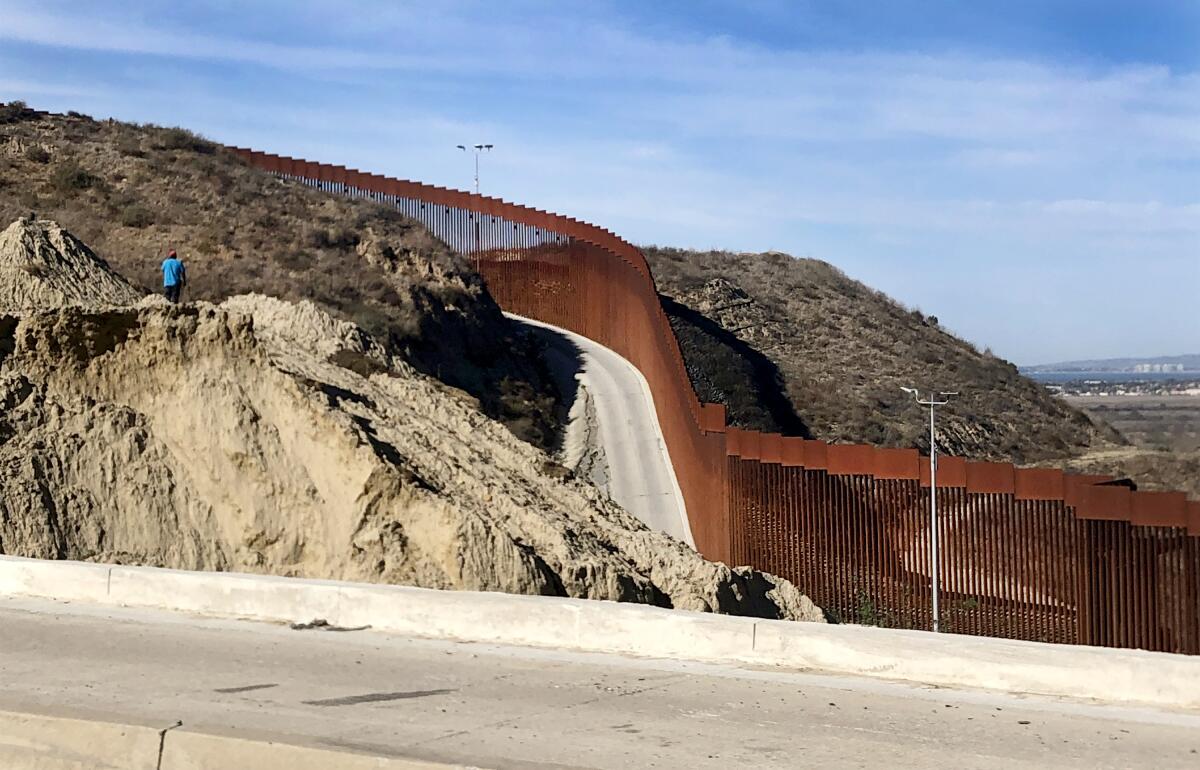 A lone man in a blue shirt, standing on the outskirts of Tijuana, Mexico, looks at a section of border wall.
