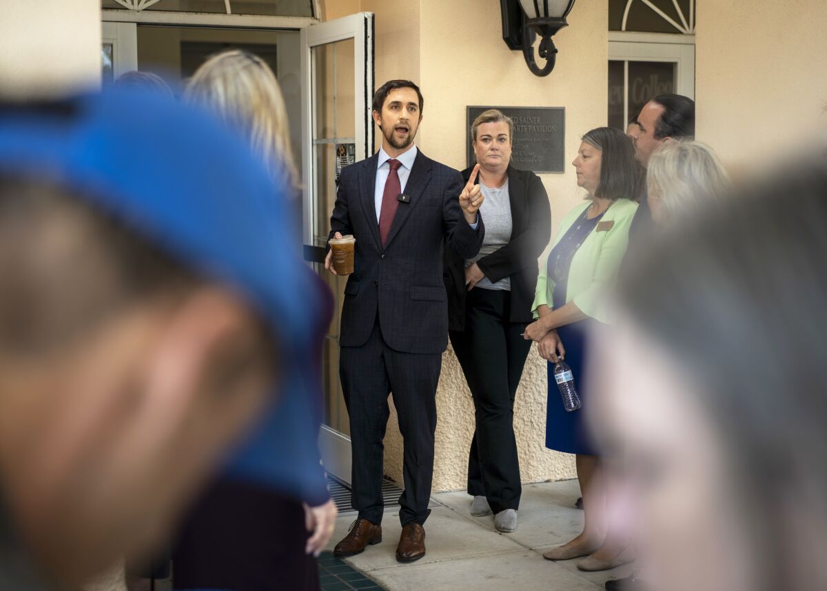 A man with dark hair and in a suit stands amid people, gestures with one hand and holds a coffee in the other.