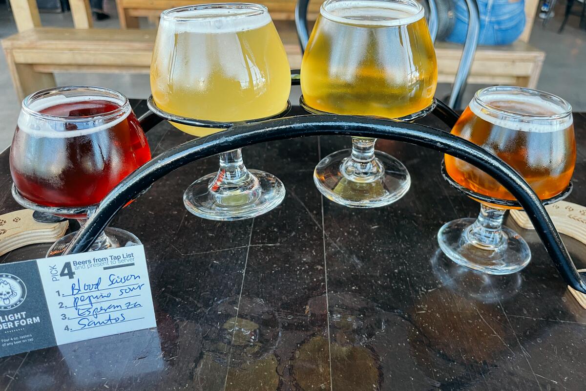A flight of four beers in footed glasses in a metal holder on a wooden table