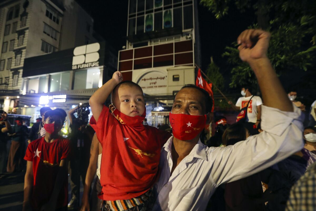 A man in a mask, carrying a child, raises his arm as supporters cheer in front of Aung San Suu Kyi's party headquarters.