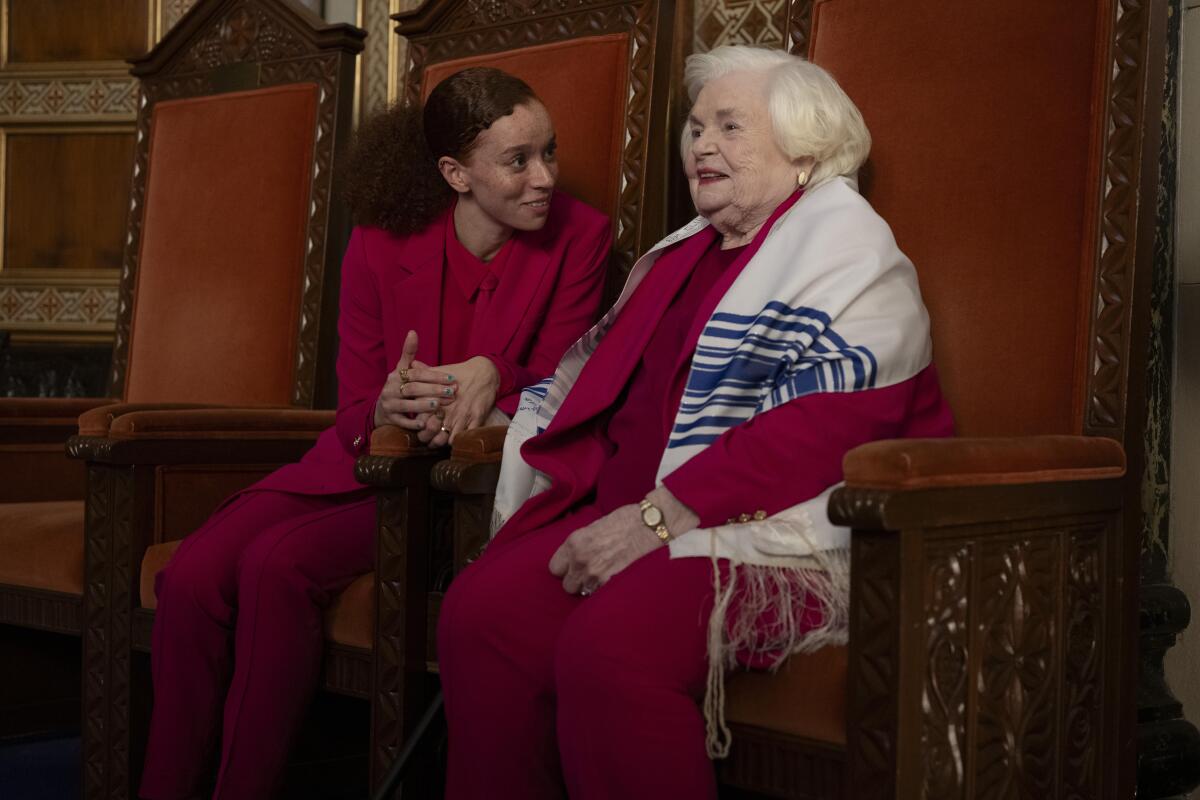 An older woman in a tallis sits in a synagogue with a friend.