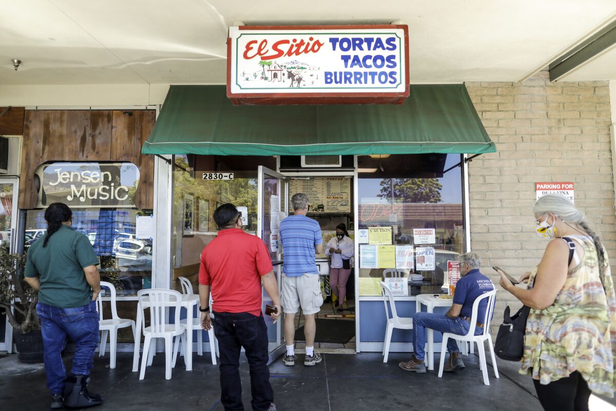 Customers wait in line to order at a small taco stand surrounded by white plastic chairs and tables