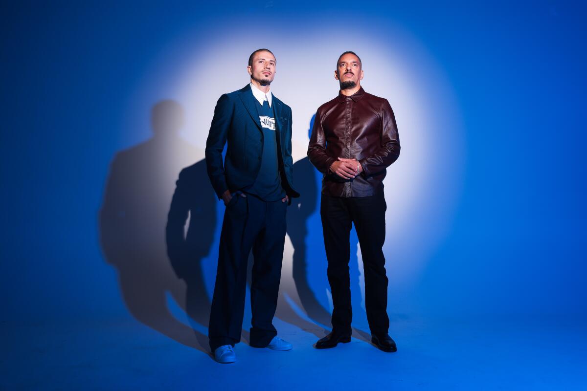 Two men stand next to each other with a spotlight on them and blue aura around them.