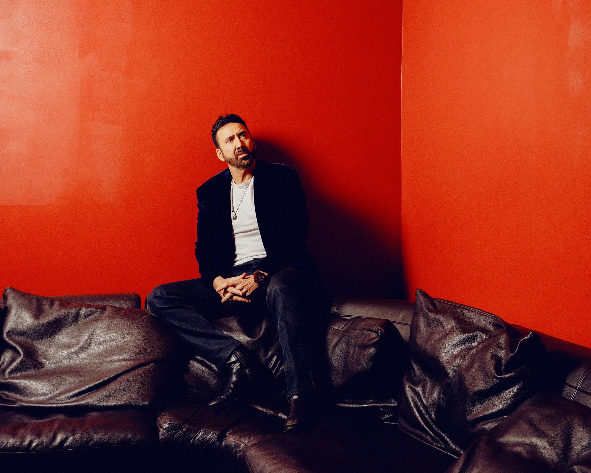 Nicolas Cage sits on the back of couch for a portrait, leaning against red walls.