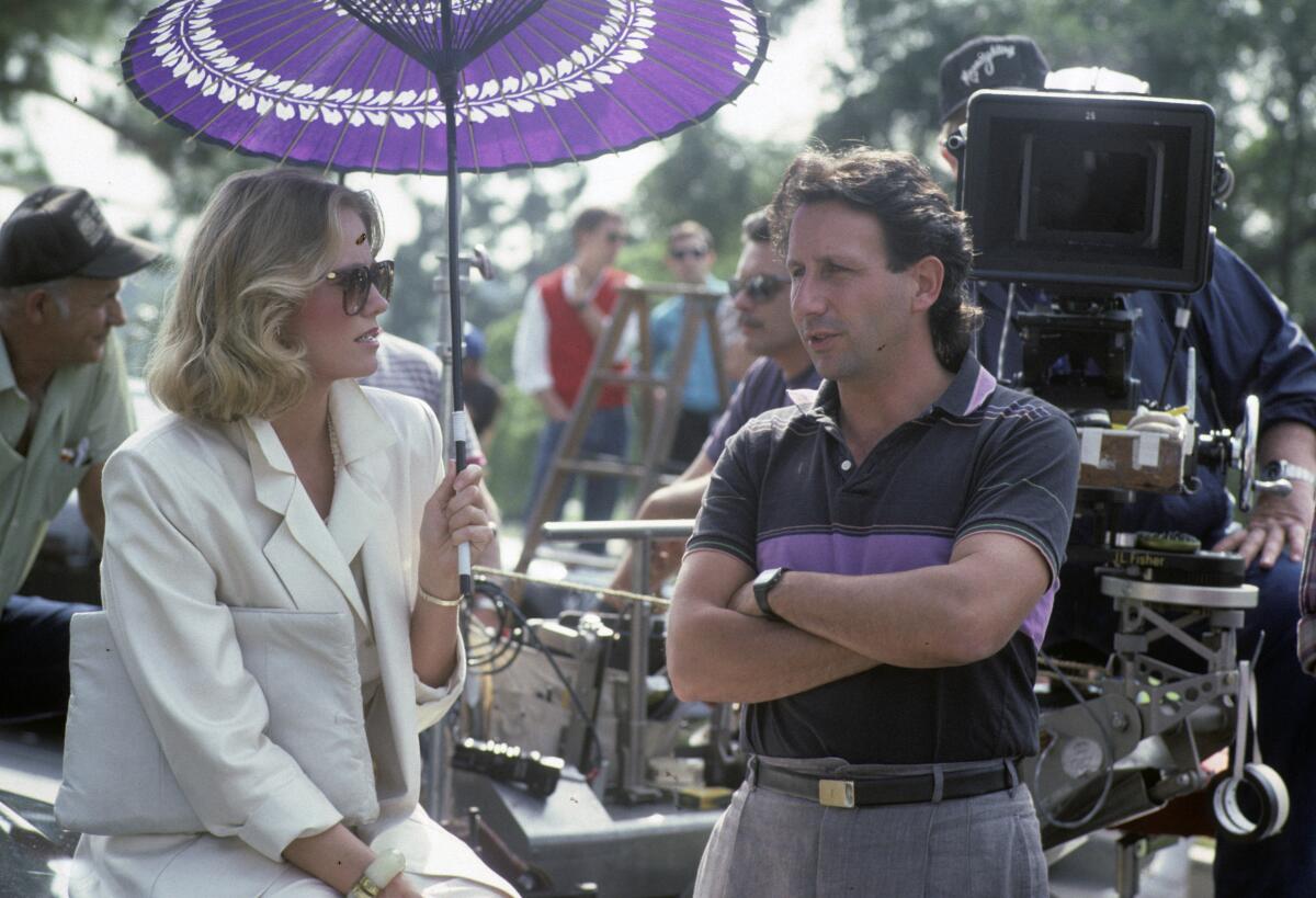 A woman with blonde hair in a white suit holds an umbrella while talking to a man standing in front of a TV camera