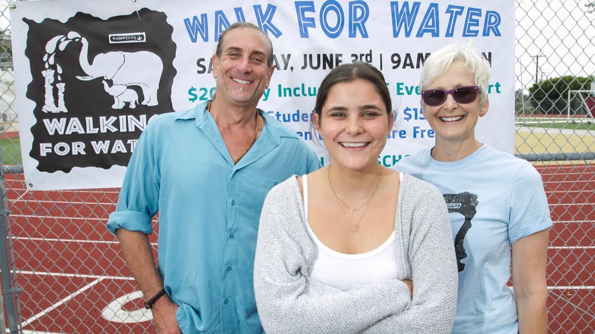 Charlotte Watkins, president of the student Walking for Water Club, center, stands with Greg Friedman, host of the Inner Journey show on KX 93.5, and Wisdom Spring board president Susan Hough in front of sign promoting Saturday's Walking for Water fundraiser.