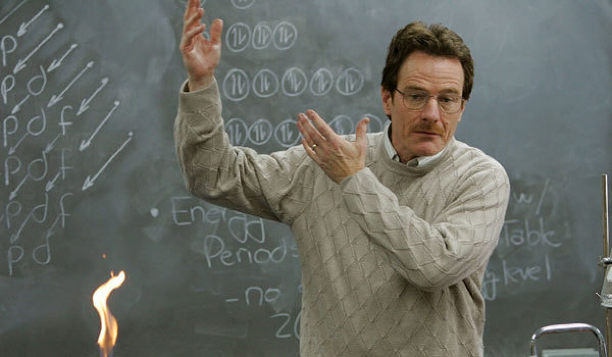 Bryan Cranston, as Walter White, gestures with both arms in front of a chalkboard. A flame burns in the foreground.