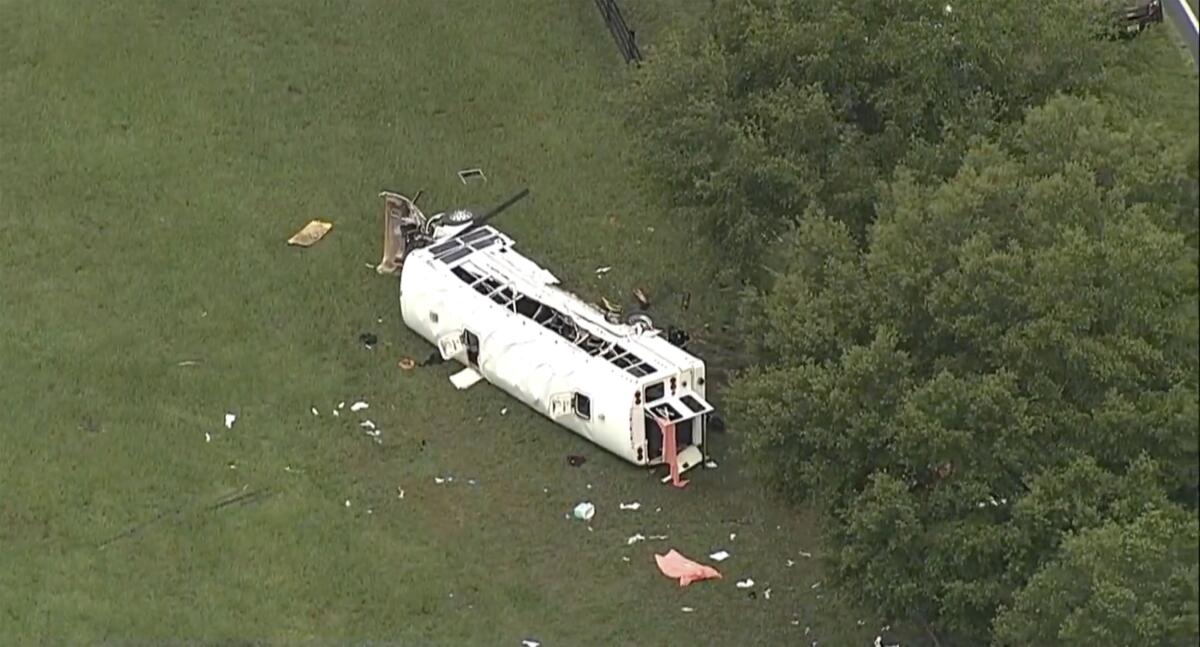 A white bus lies on its side with debris scattered around it at the edge of trees.