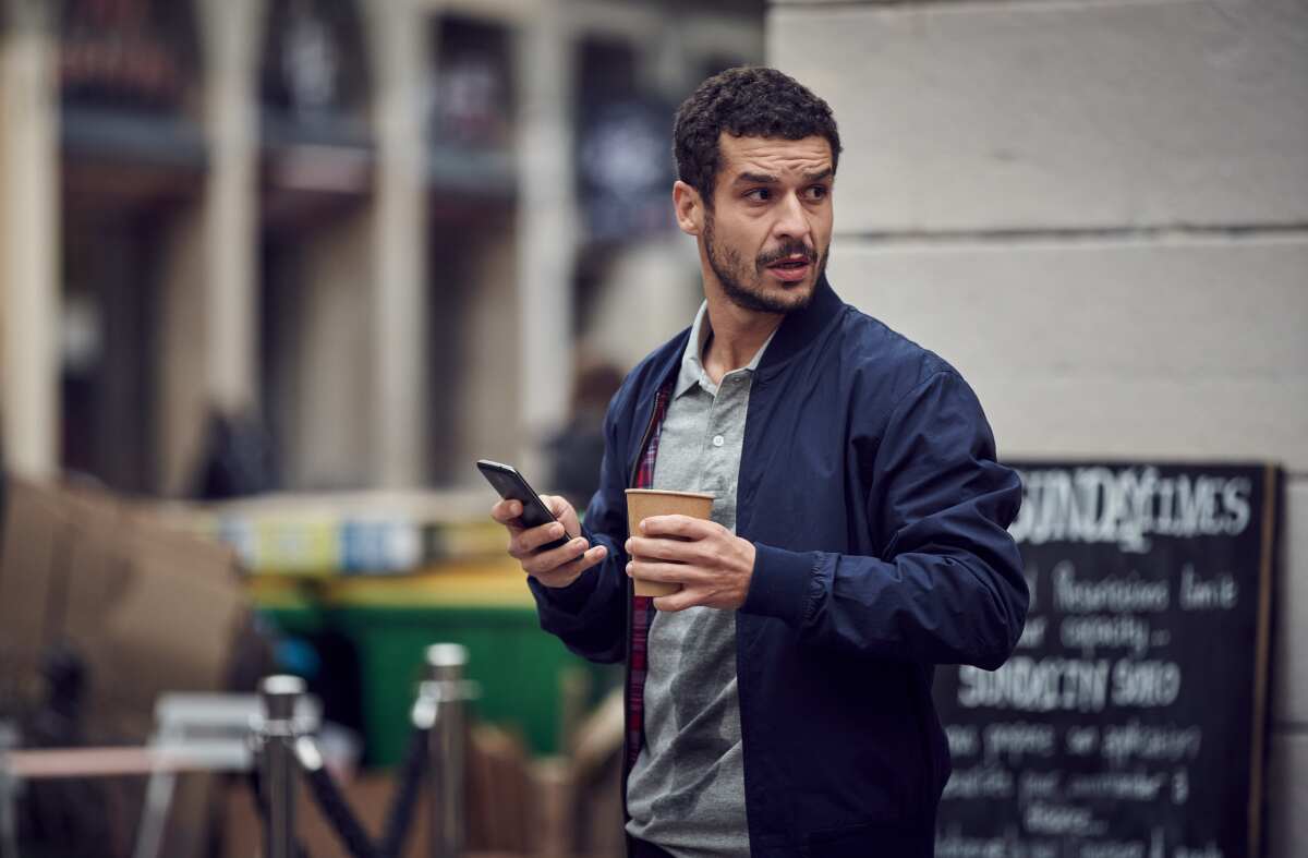 A bearded man in a windbreaker, holding a cellphone and a to-go coffee cup, looks to the side with a puzzled expression.