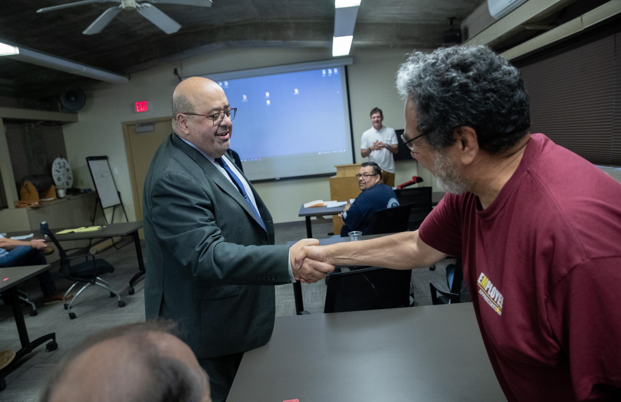 Adel Hagekhalil, wearing glasses and a large gray suit, shakes hands with a man wearing a red T shirt while at a presentation