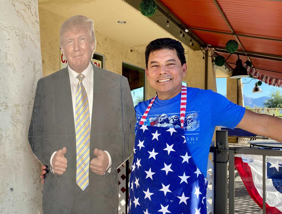 Jorge Rivas wears a red white and blue apron and smiles next to a cardboard cutout of President Trump