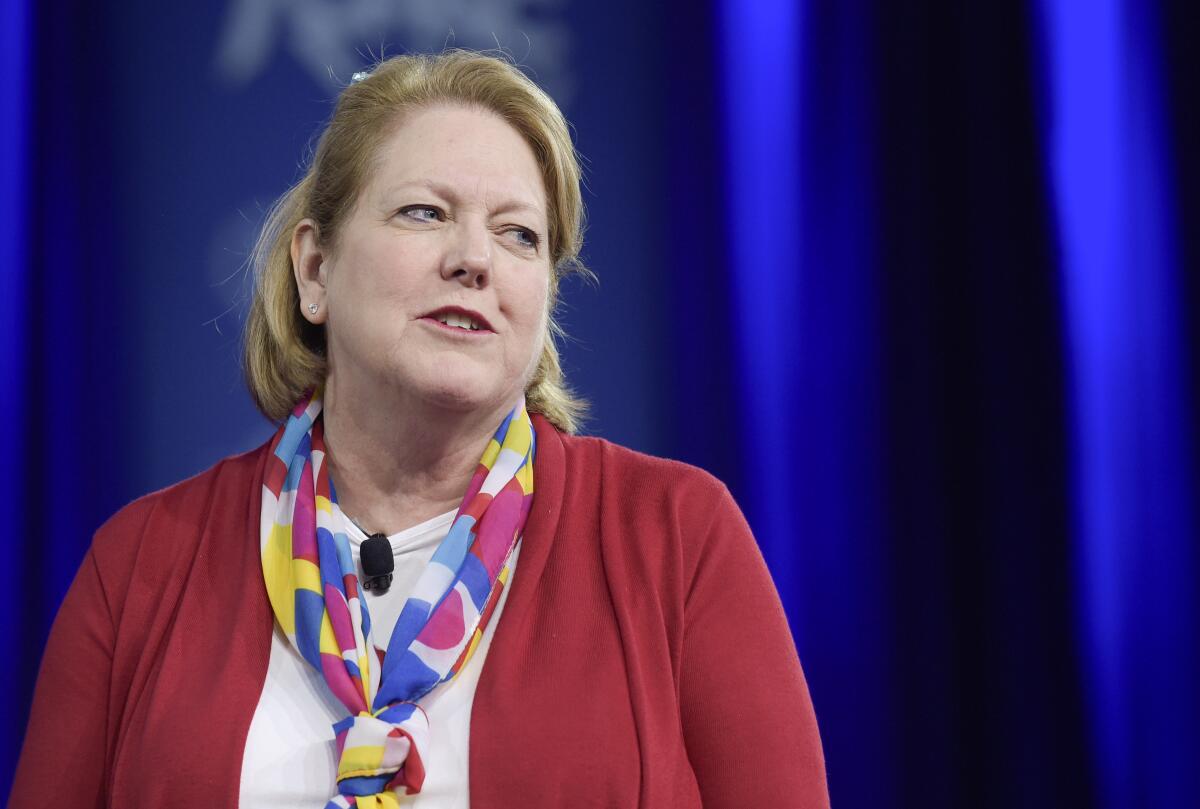 A woman with blond hair, in a red jacket, white top and colorful scarf, looks to the right 