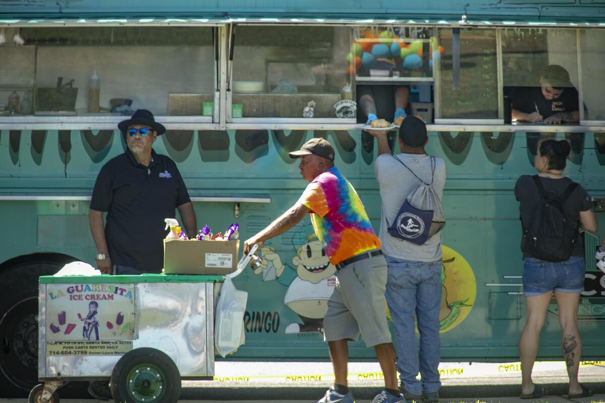 An ice cream vendor manages his push cart as a taco truck serves customers.