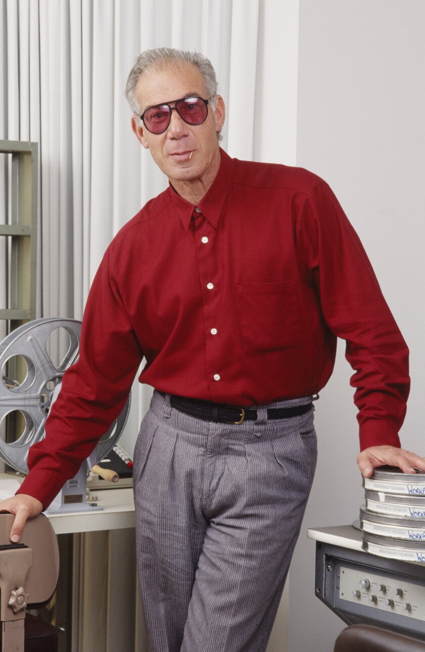 A man posing in sunglasses, pants and a button-up shirt.