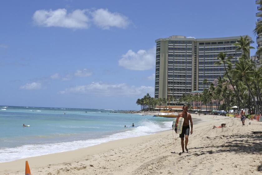 In this photo taken Friday, June 5, 2020, a surfer walks on a sparsely populated Waikiki beach in Honolulu. Hawaii faces unpleasant options for addressing a dramatic decline in tax revenues precipitated by the coronavirus pandemic and the shutdown of the state's tourism industry. (AP Photo/Audrey McAvoy)