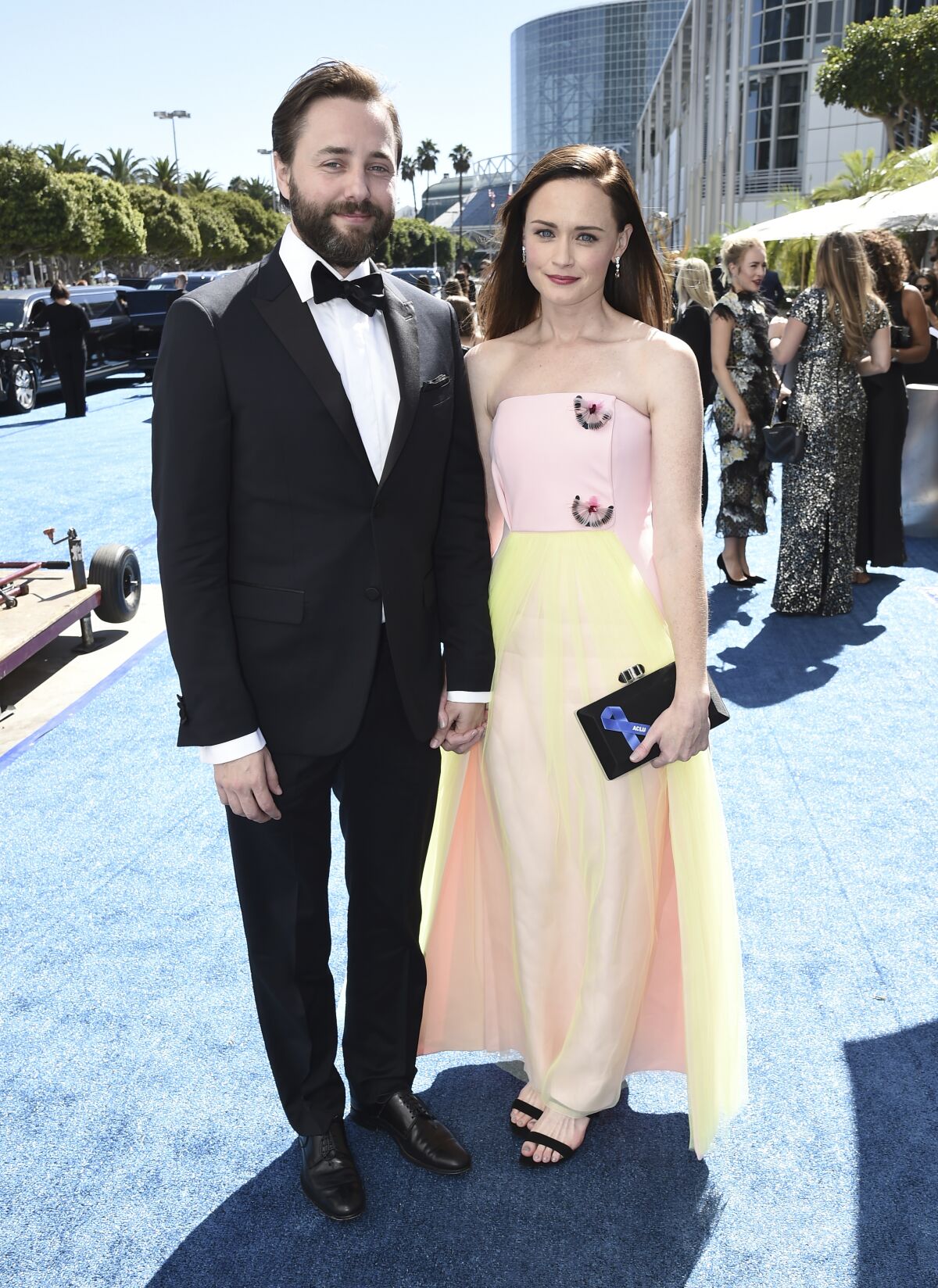 A bearded man in a tuxedo stands next to a woman wearing a pink and orange gown on a blue carpet.
