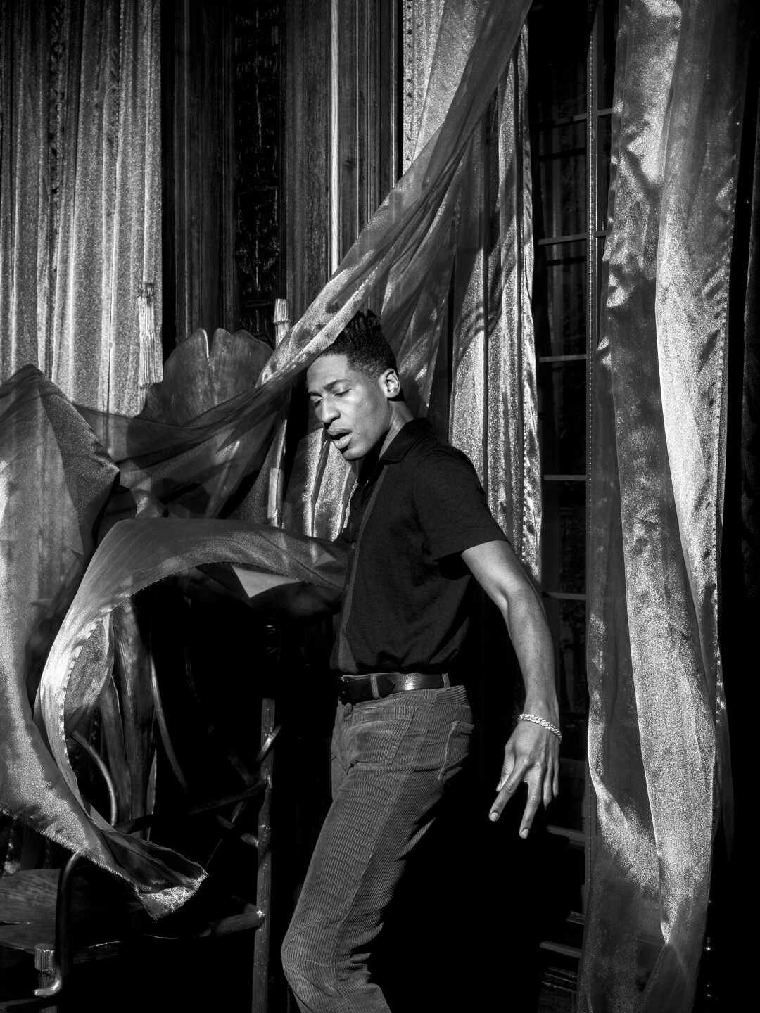 A man in a dark shirt and gold chain dances with sheer curtains.