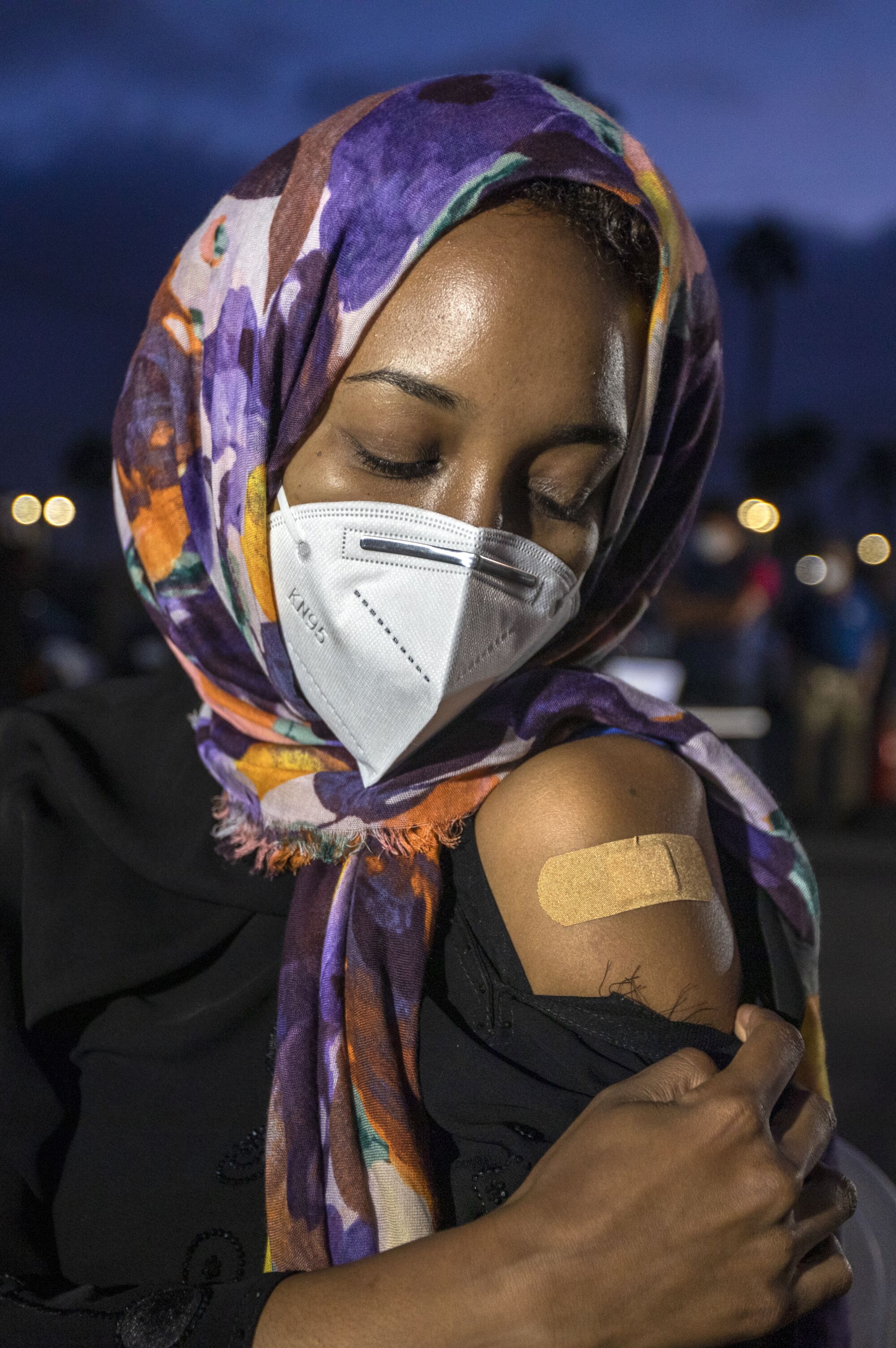 A woman in a headscarf and mask shows a Band-Aid on her upper arm.