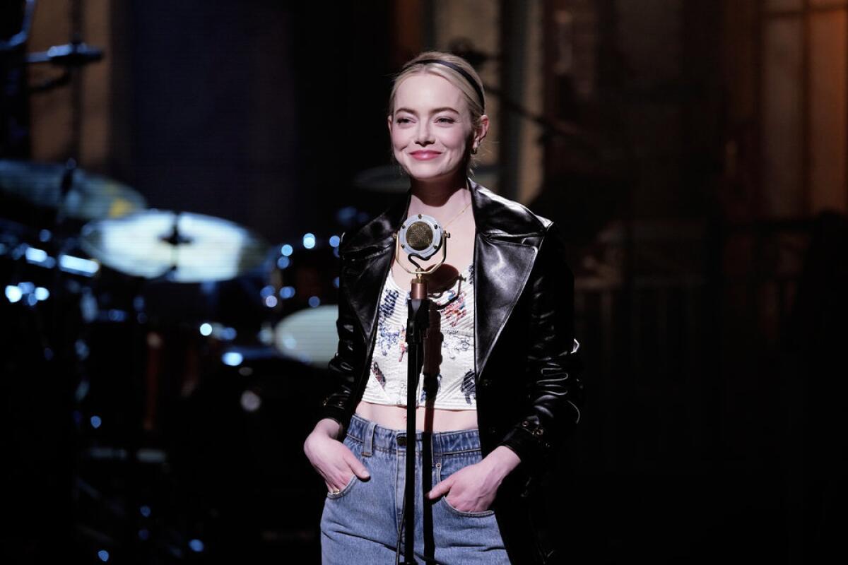 Emma Stone stands on a stage in front of a microphone with her hands in her jean pockets in a black jacket and white top.