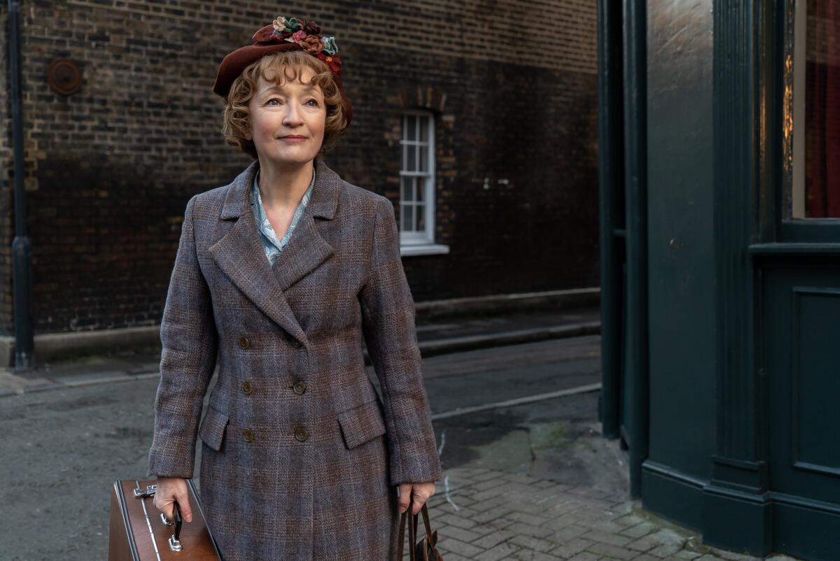 A women in a coat and hat and holding a suitcase stands in a street and smiles.