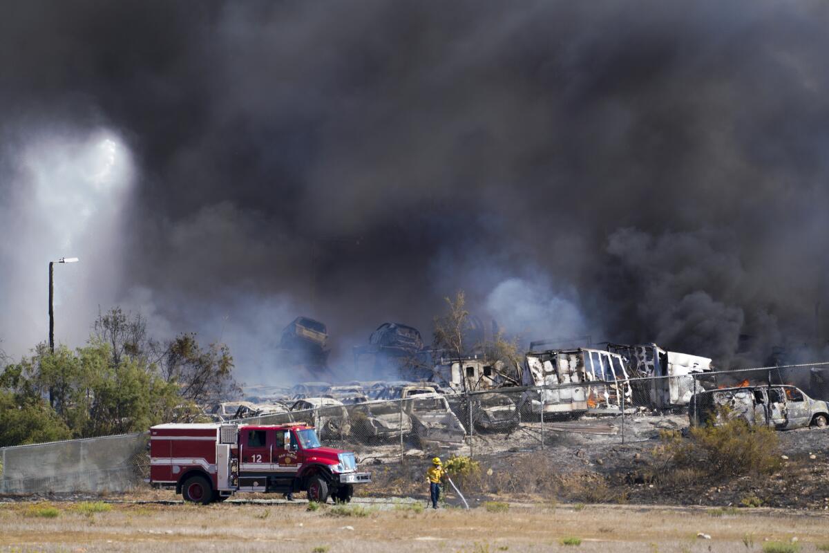 Firefighters work to extinguish an inferno at a wrecking yard while black smoke fills the sky.